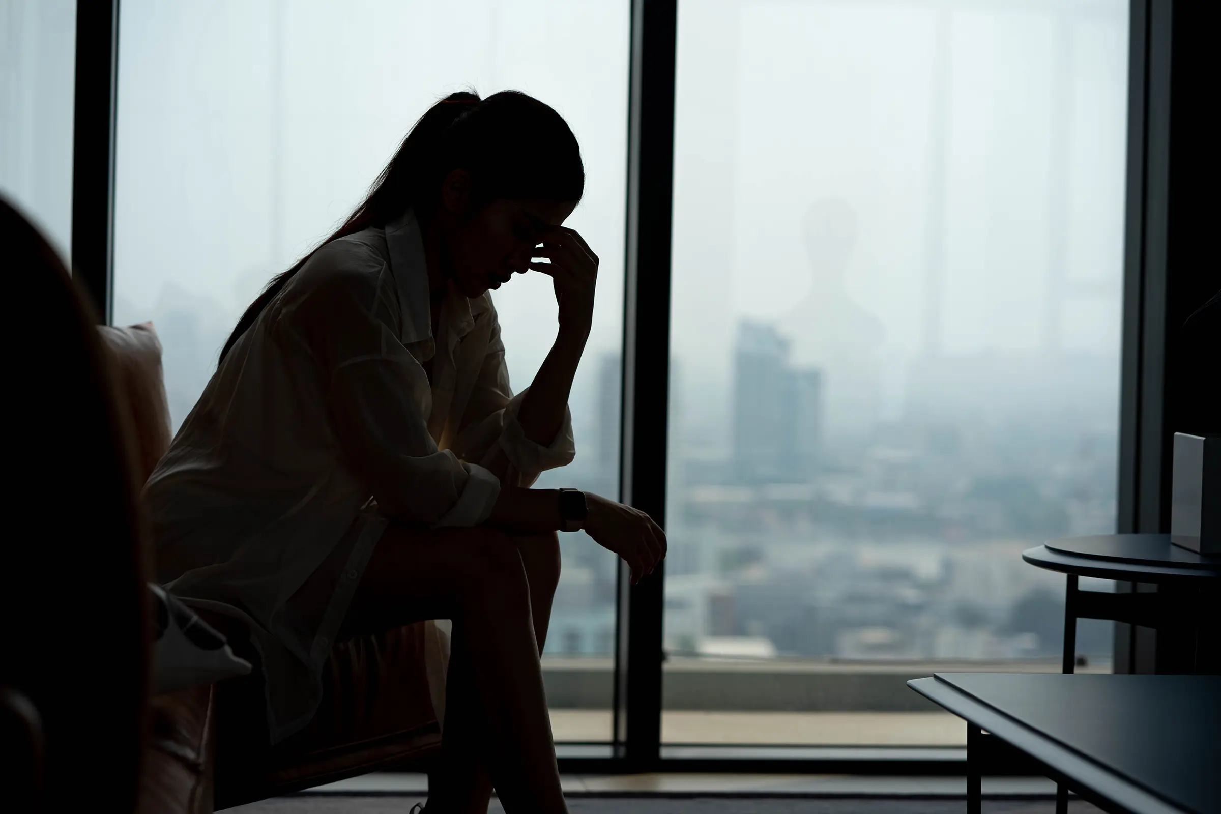 A woman sits alone in silhouette against an office window, head lowered and hand raised to her face.
